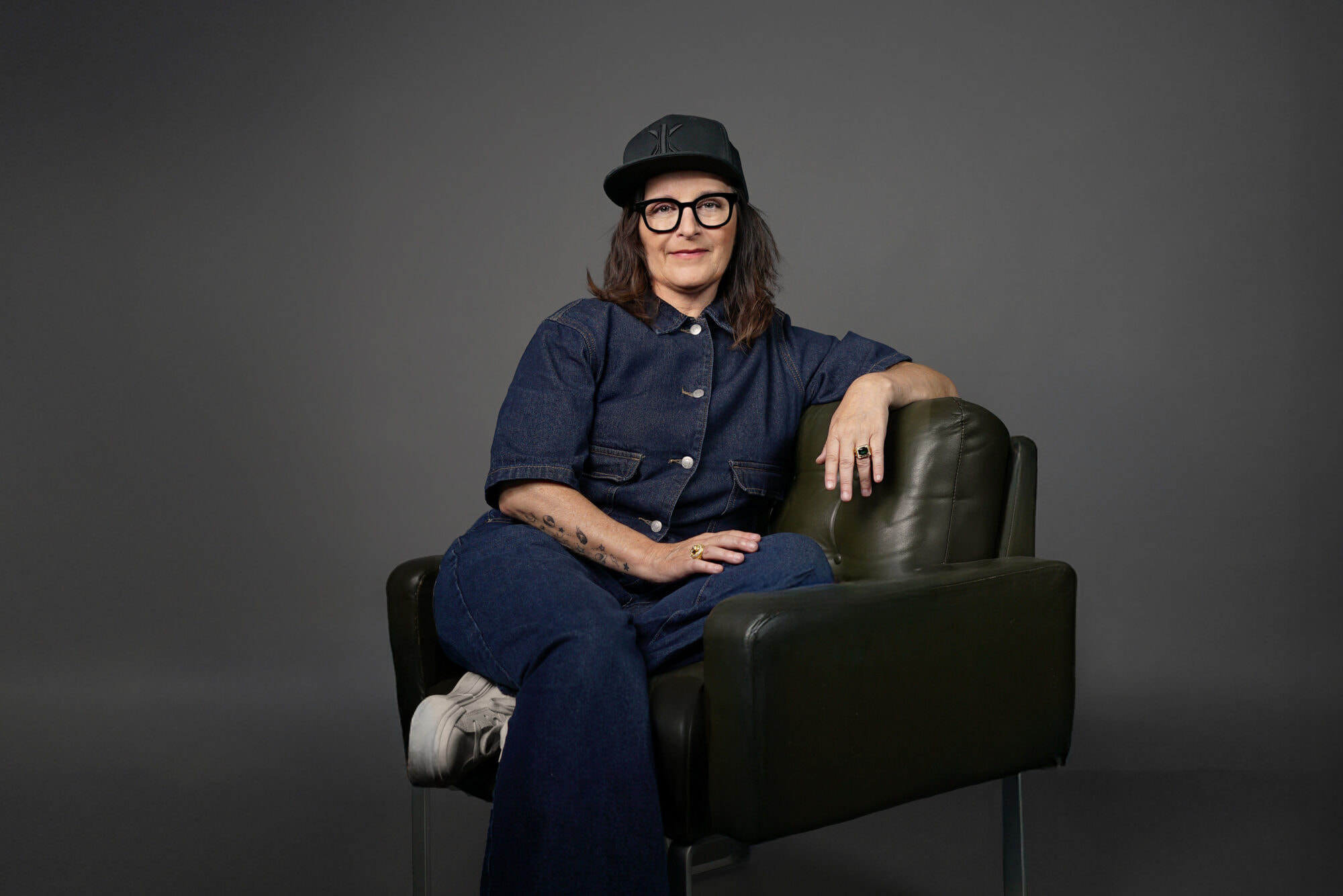 Bild von Anja Maria Welzel wie sie sitzend auf einem Sessel in die Kamera guckt. Sie trägt ein Cap und eine Brille.