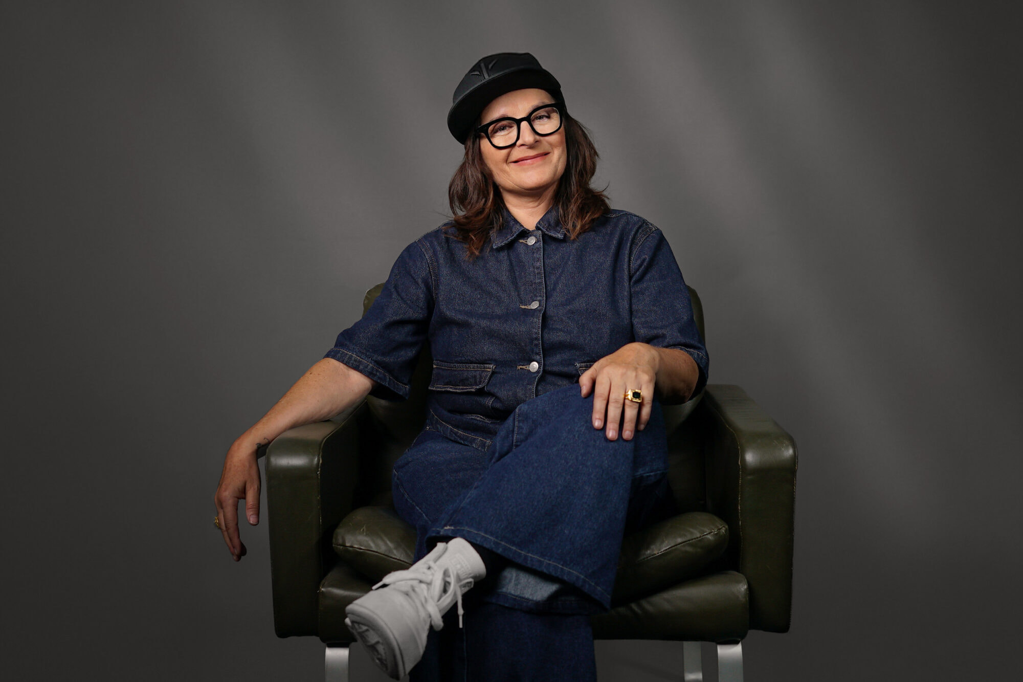 Bild von Anja Maria Welzel wie sie sitzend auf einem Sessel in die Kamera guckt. Sie trägt ein Cap und eine Brille.