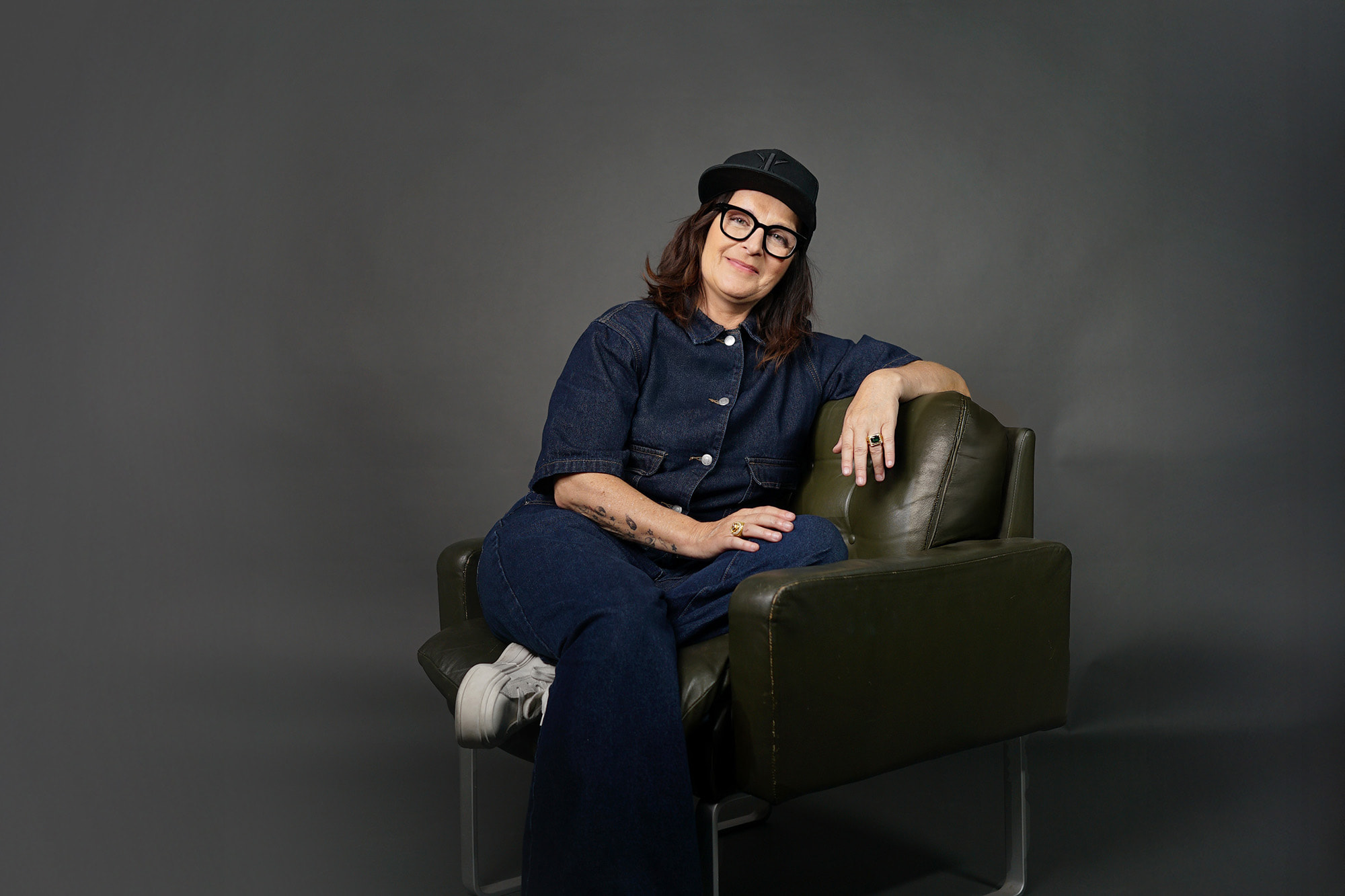 Bild von Anja Maria Welzel wie sie sitzend auf einem Sessel in die Kamera guckt. Sie trägt einen Jeans Anzug, ein Cap und eine Brille.